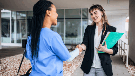 woman in scrubs shaking hands with woman in casual business attire holding clipboard
