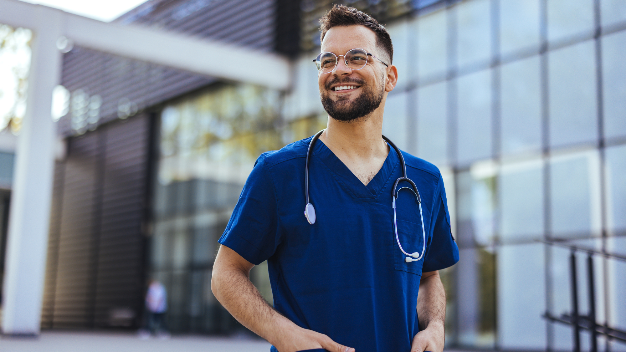 Male nurse in blue scrubs standing in front of facility, smiling - healthcare recruitment strategies/finding the perfect candidate