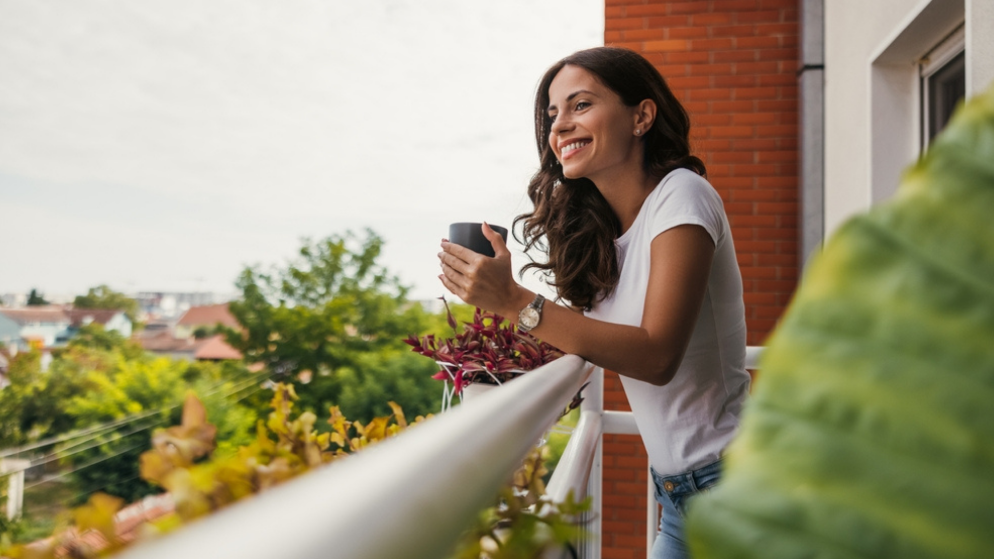 Young woman standing on an apartment balcony holding a cup of coffee.