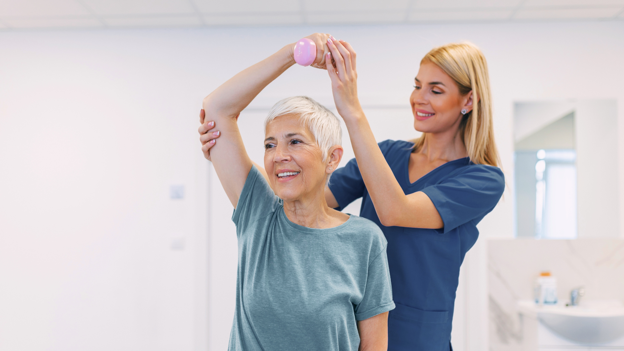 Blonde woman helping elderly woman lift weight in PT