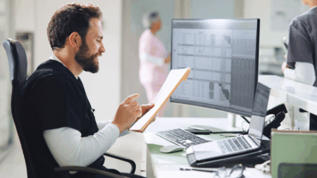 a-smiling-man-in-scrubs-sitting-at-computer-looking-at-folder-in-hand