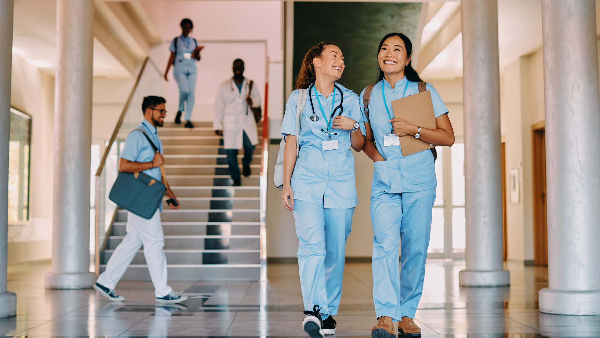 Two female nurses wearing blue scrubs smiling with each other as they walk down the hall of a hospital