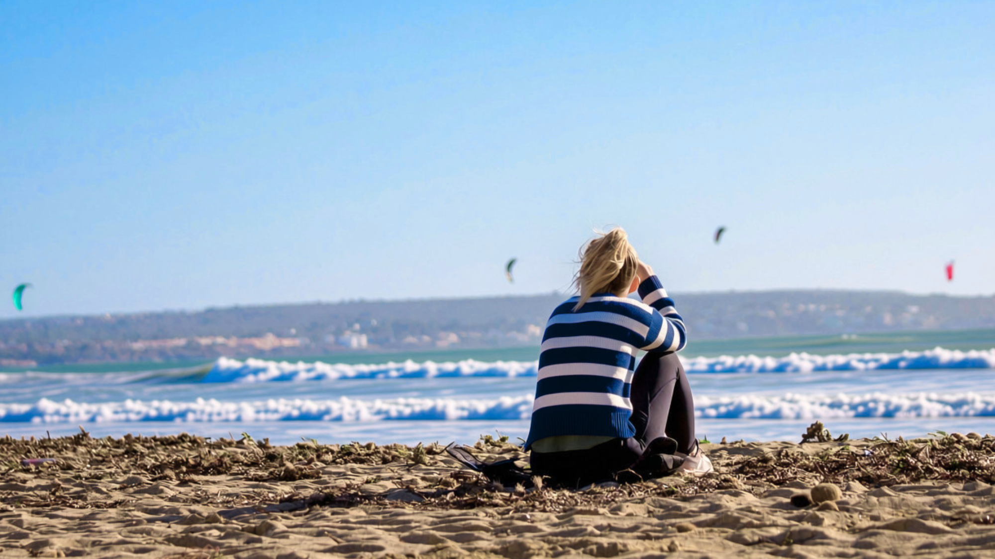 Woman in blue and white striped sweater sitting on the beach on a sunny day and staring at the ocean.