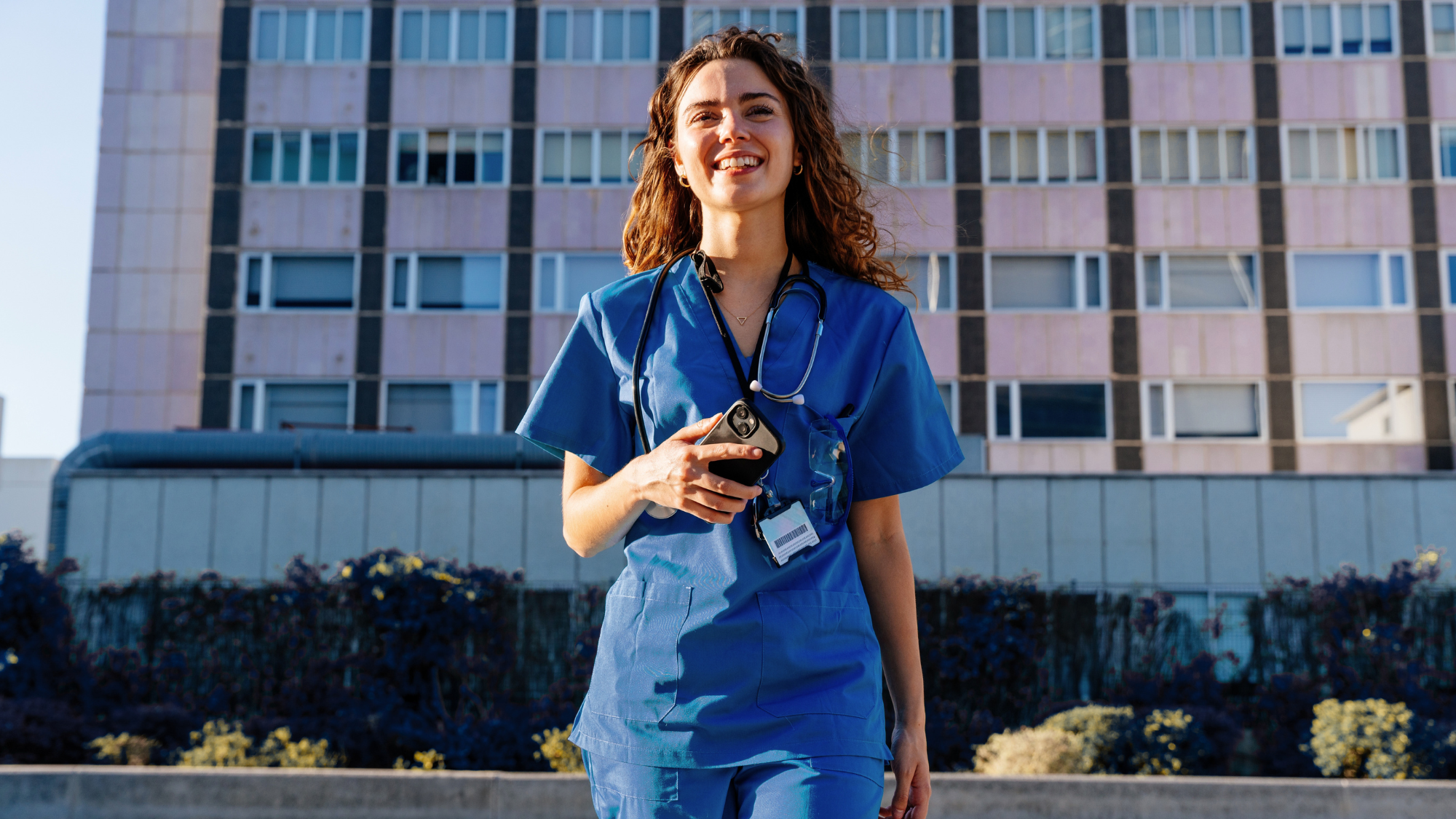 Female nurse standing in front of a hospital smiling holding her phone.