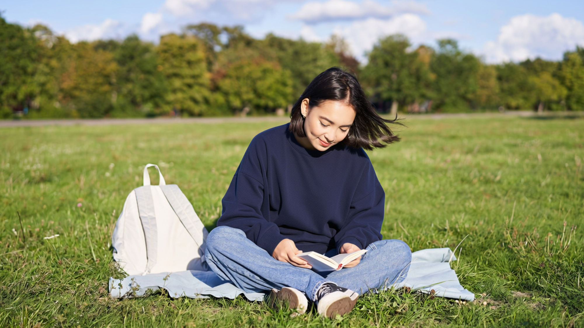 Woman sitting on a blanket in a park reading a book.