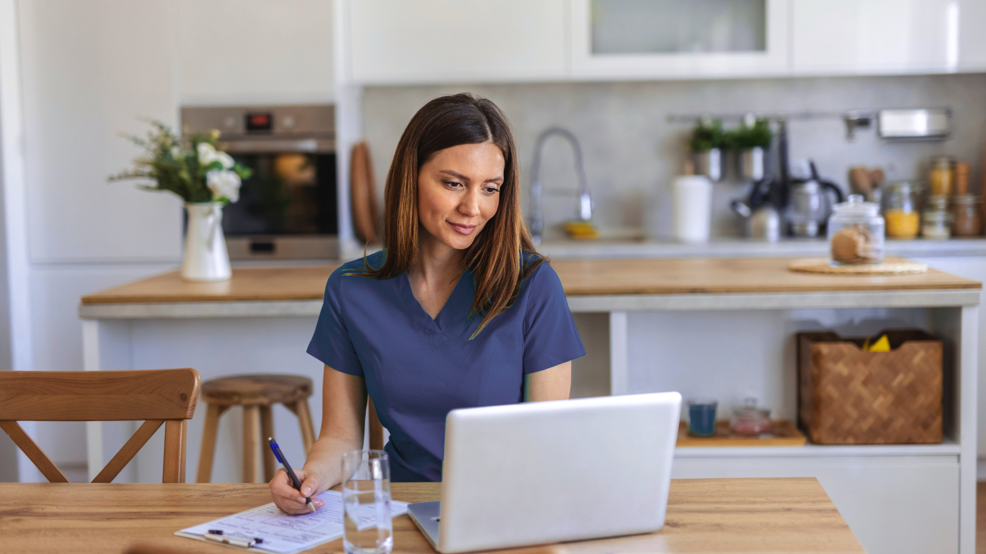 Female nurse with brown hair and navy scrubs writing on paper while looking at a laptop in a kitchen.