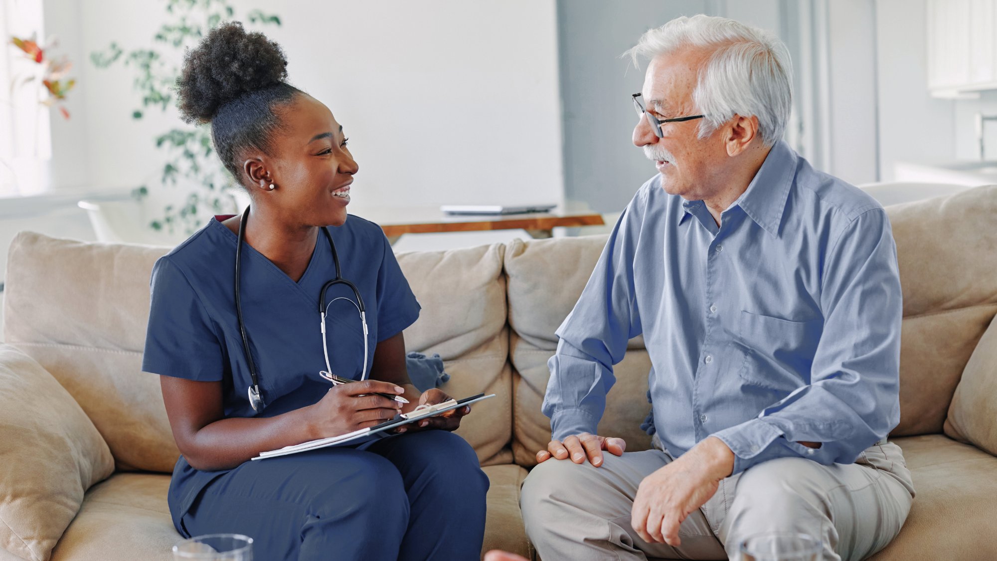 A female nurse sitting with an elderly man on a couch talking.
