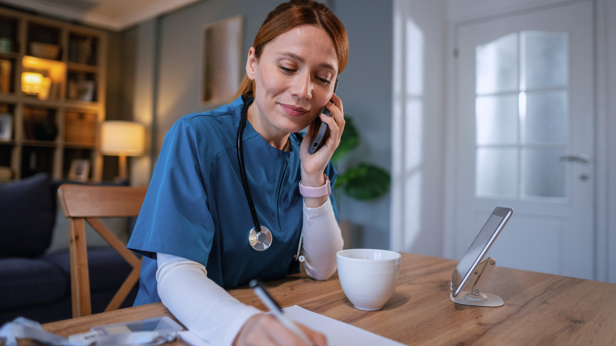 A nurse on the phone sitting at table writing while on a travel nursing assignment.